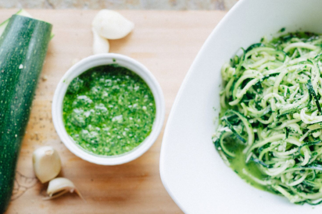 One whole green zucchini sits on a cutting board next to a bowl of spiralized zucchini noodles or zoodles. Another bowl with green pesto sits waiting to be poured into the noodles.
