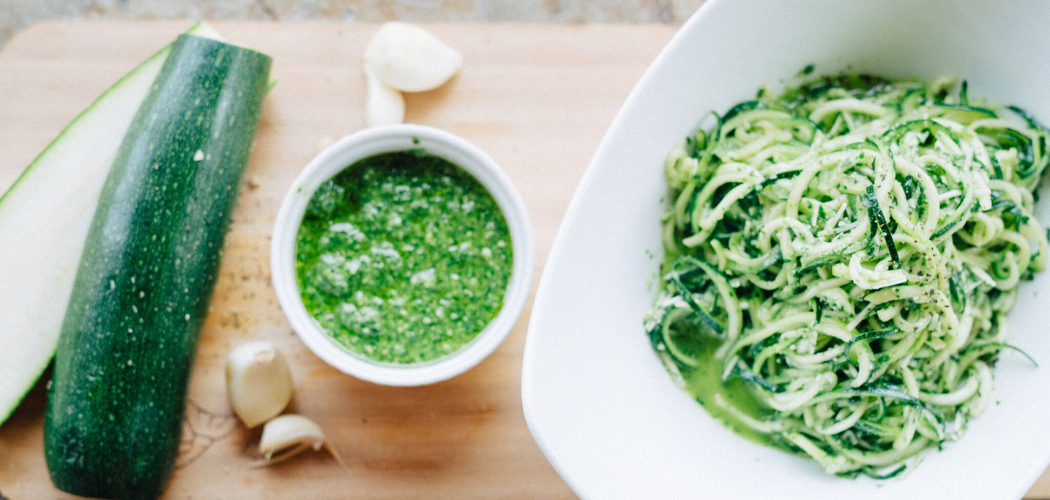 One whole green zucchini sits on a cutting board next to a bowl of spiralized zucchini noodles or zoodles. Another bowl with green pesto sits waiting to be poured into the noodles.