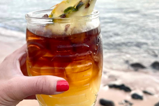 Nealy Fisher holds a skinny Mai Tai with a pineapple as garnish on top in front of the shore in Hawaii.