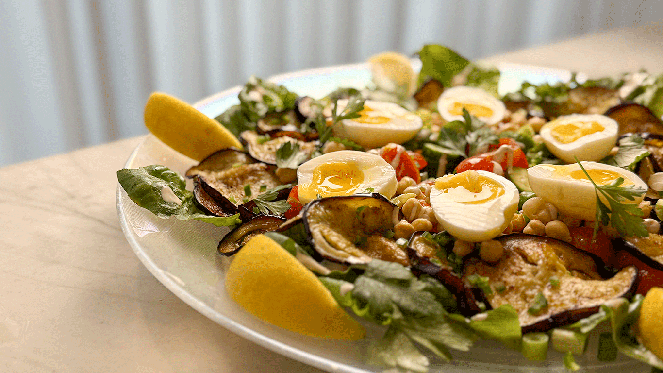 Colorful Sabich Salad with soft-boiled eggs, eggplant slices, chickpeas, lettuce, and lemon wedges on a glass plate