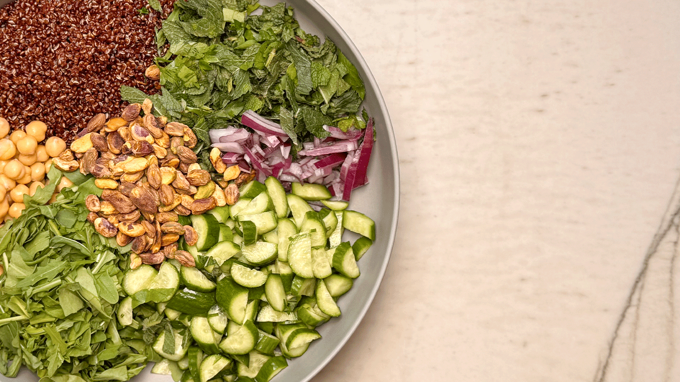 Bowl of ingredients for quinoa salad with chickpeas, pistachios, cucumbers, herbs, red onion, arugula, and red quinoa