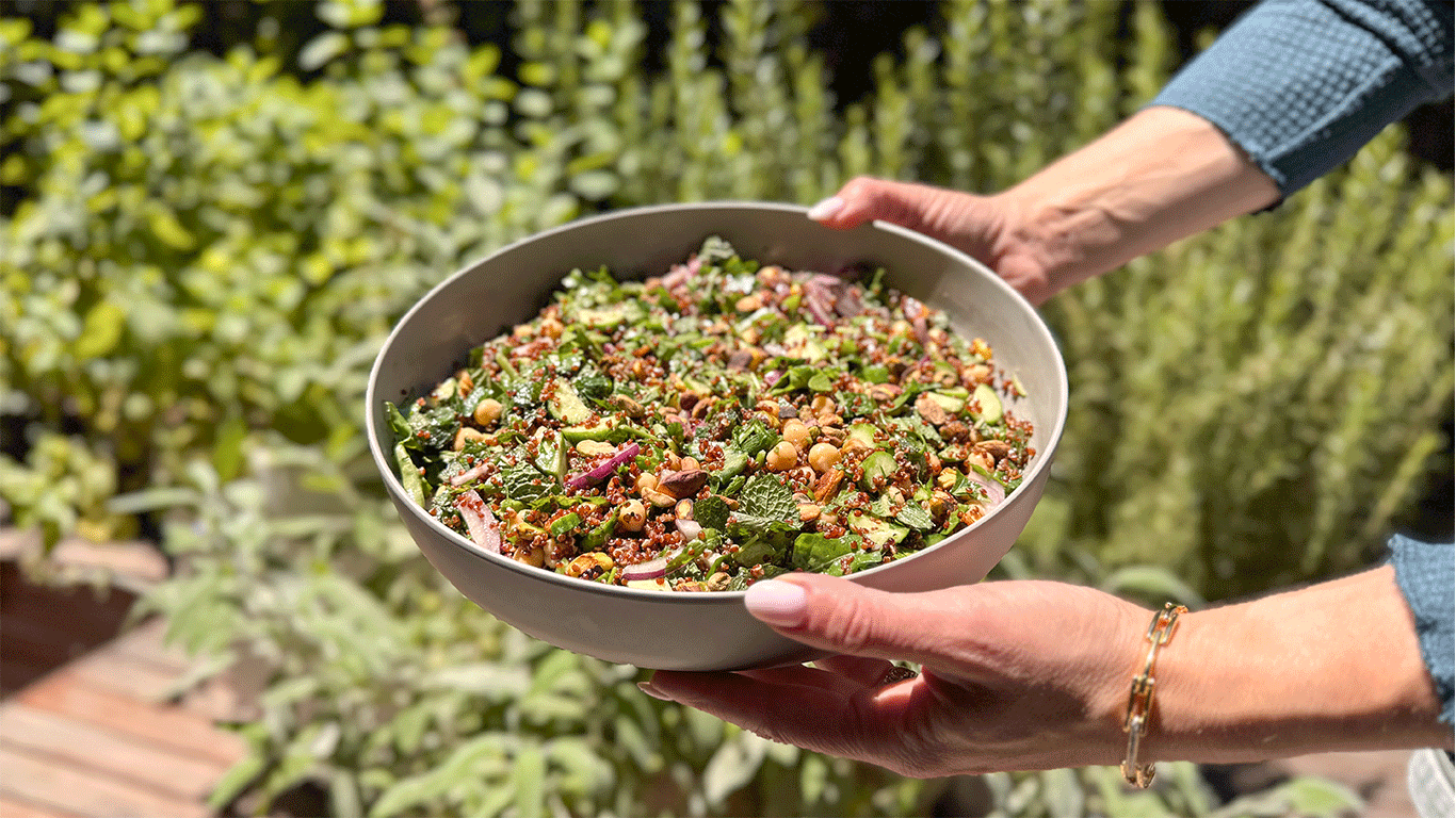 A bowl of quinoa salad with chickpeas, pistachios, cucumbers, and herbs being held outdoors in natural light