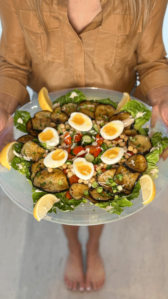 Nealy Person holding a glass plate of Sabich Salad with eggplant, soft-boiled eggs, chickpeas, lettuce, tomatoes, and lemon wedges