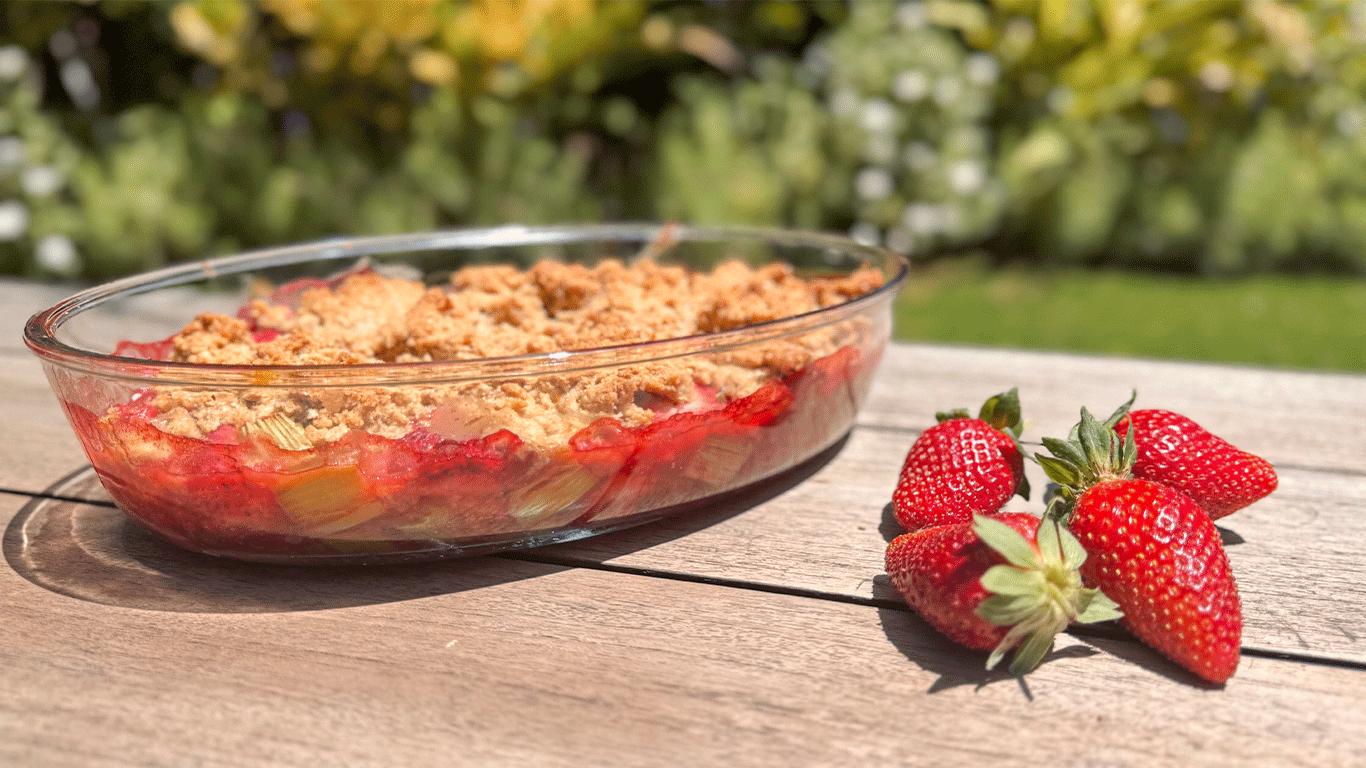 Strawberry rhubarb crumble in a glass pie dish with fresh strawberries on a wooden table outdoors – gluten-free dessert with fruit and crumble topping