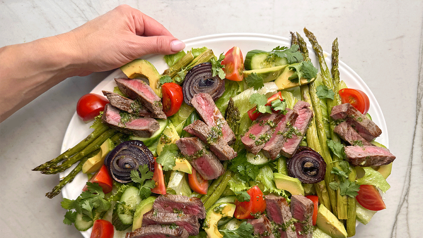 Overhead view of a colorful steak salad with grilled asparagus, red onion, avocado, tomatoes, cucumbers, and chimichurri dressing.