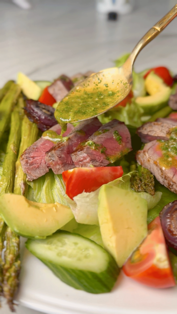Close-up of chimichurri sauce being spooned over slices of seared steak on a colorful vegetable salad.