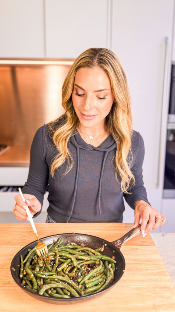 Woman in a dark hoodie holds a forkful of sautéed green beans in a skillet, ready to enjoy a simple, healthy meal.
