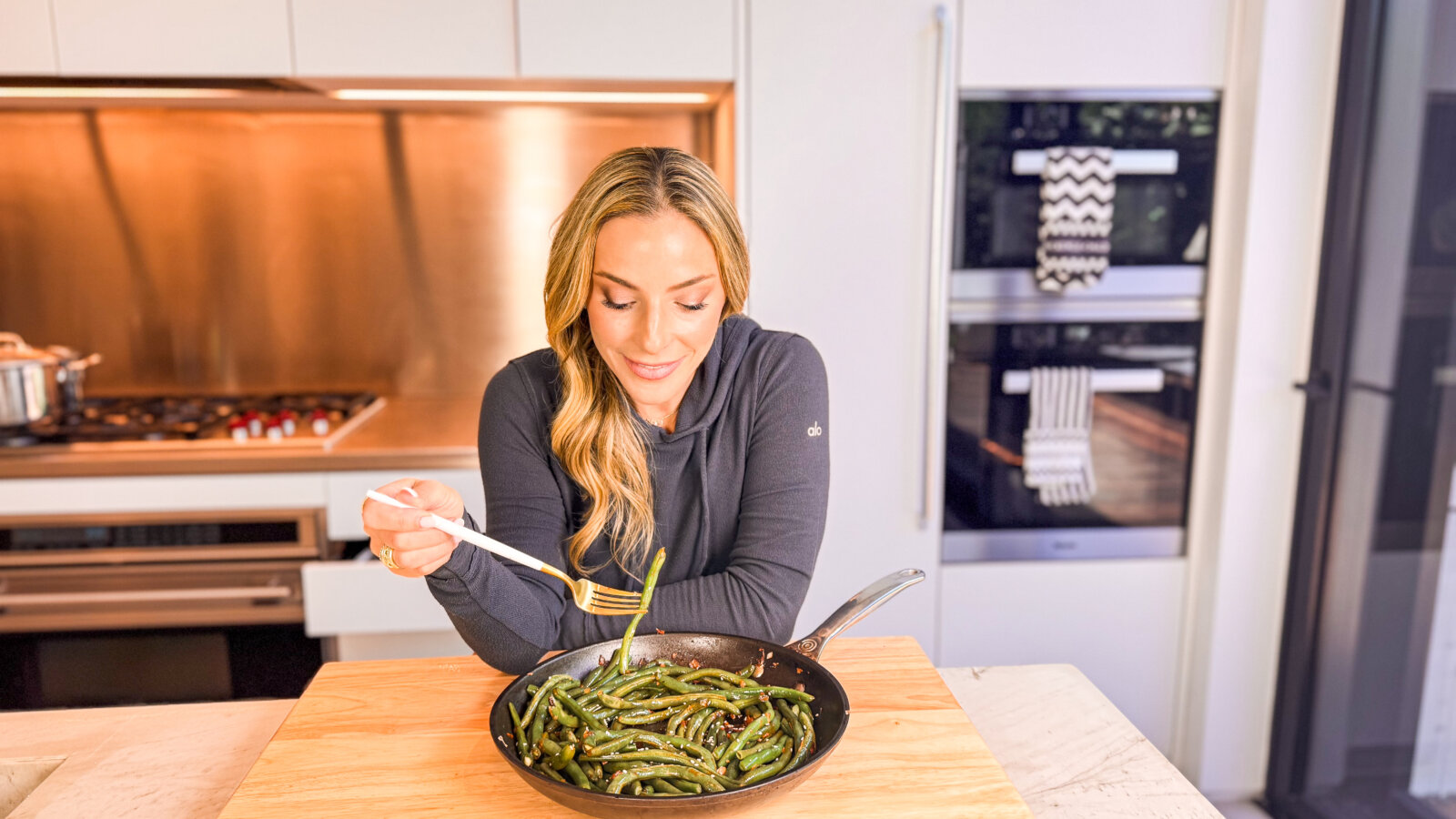 A woman in a cozy kitchen smiles down at a skillet filled with sautéed green beans, ready to take a bite with a gold and white fork.