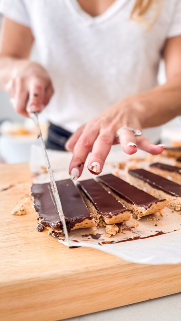 Close-up of hands slicing chocolate peanut butter bars on a wooden board with a knife, showing rich layers of chocolate and nutty filling.