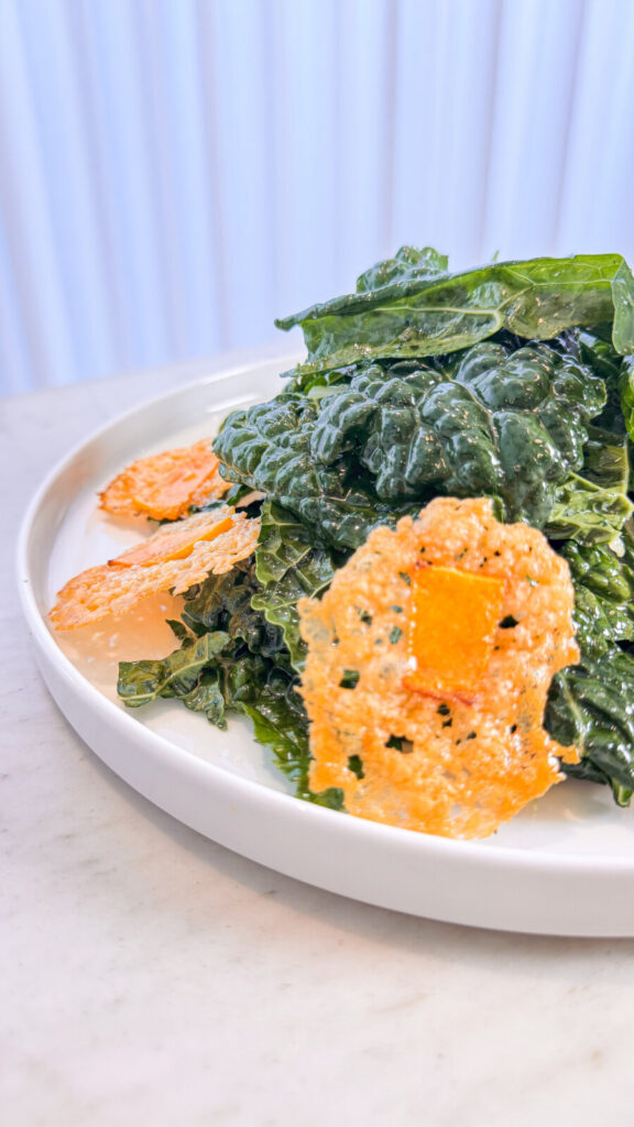 Close-up of a kale salad topped with crisp pumpkin parmesan croutons on a white plate over a marble surface.
