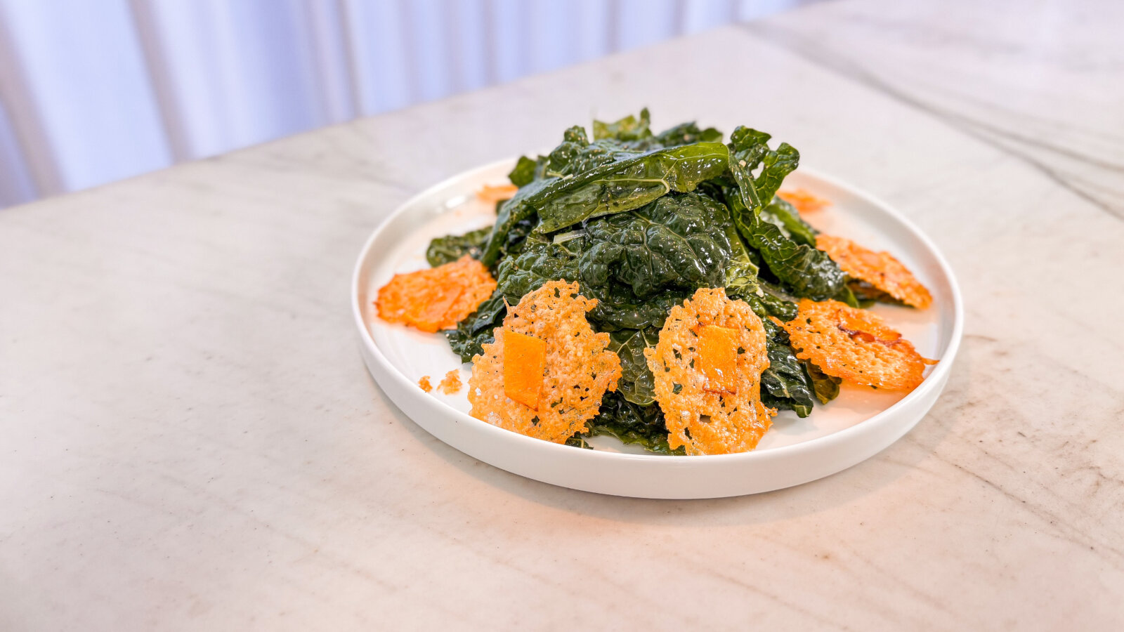 A plate of kale salad topped with crisp pumpkin parmesan croutons, served on a white plate over a marble surface.