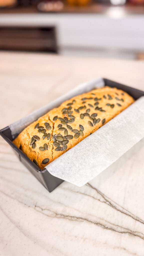 A freshly baked golden loaf topped with pumpkin seeds, resting in a parchment-lined loaf pan on a marble countertop.