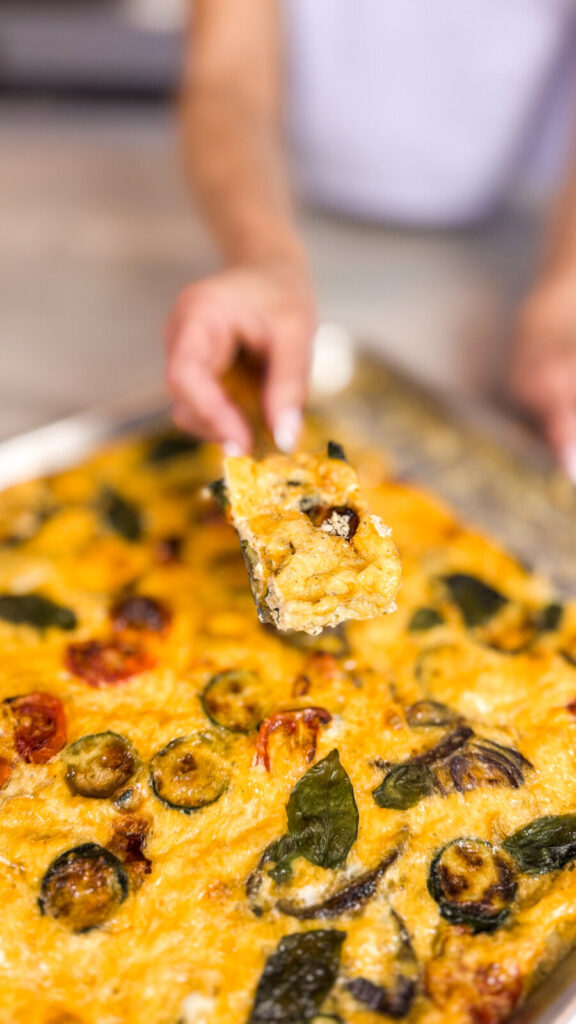 Close-up of a hand holding a slice of golden sheet pan frittata topped with roasted vegetables and herbs.