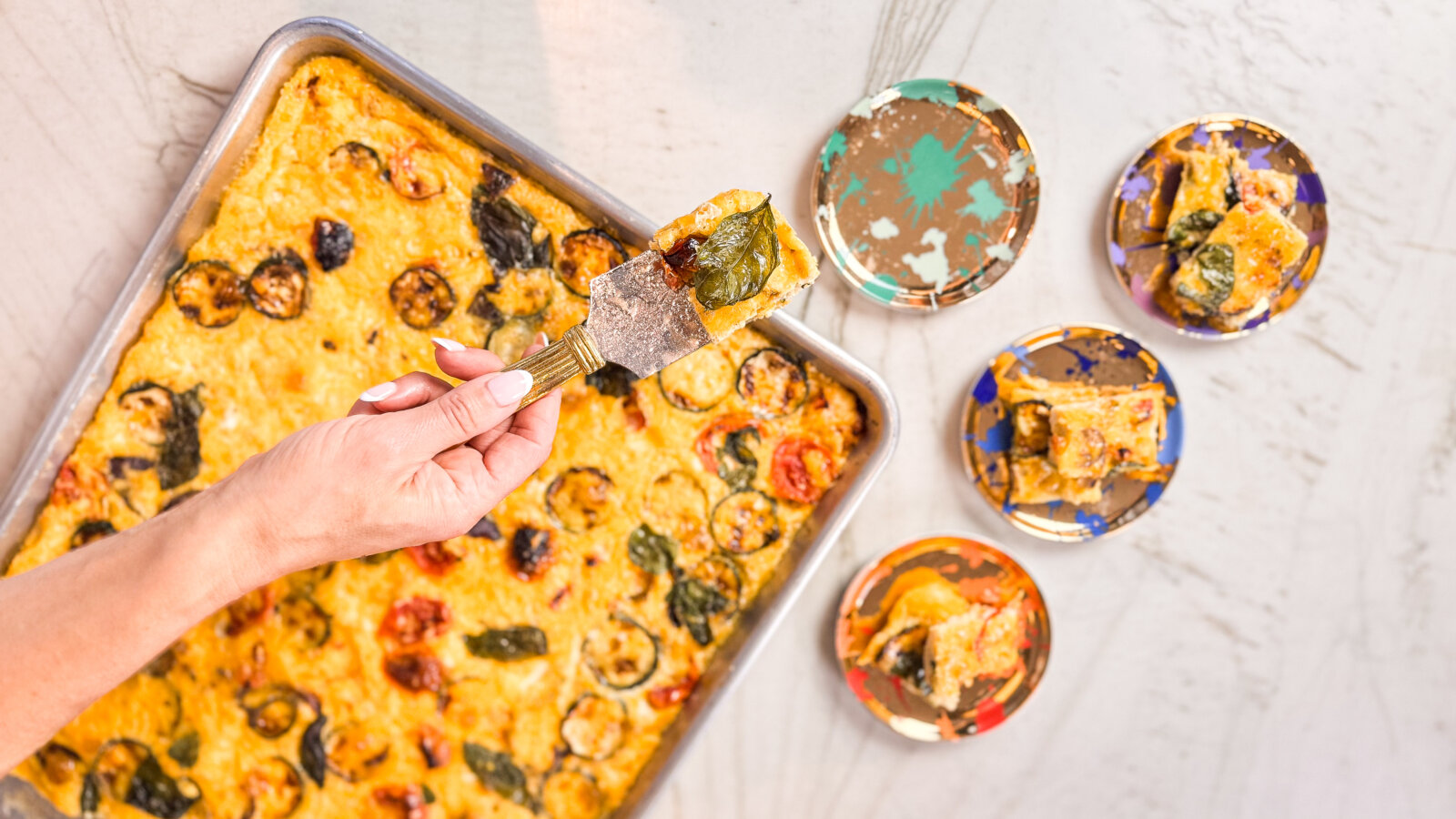 A hand lifts a slice of golden sheet pan frittata topped with roasted vegetables and herbs, with several colourful plates holding portions nearby.