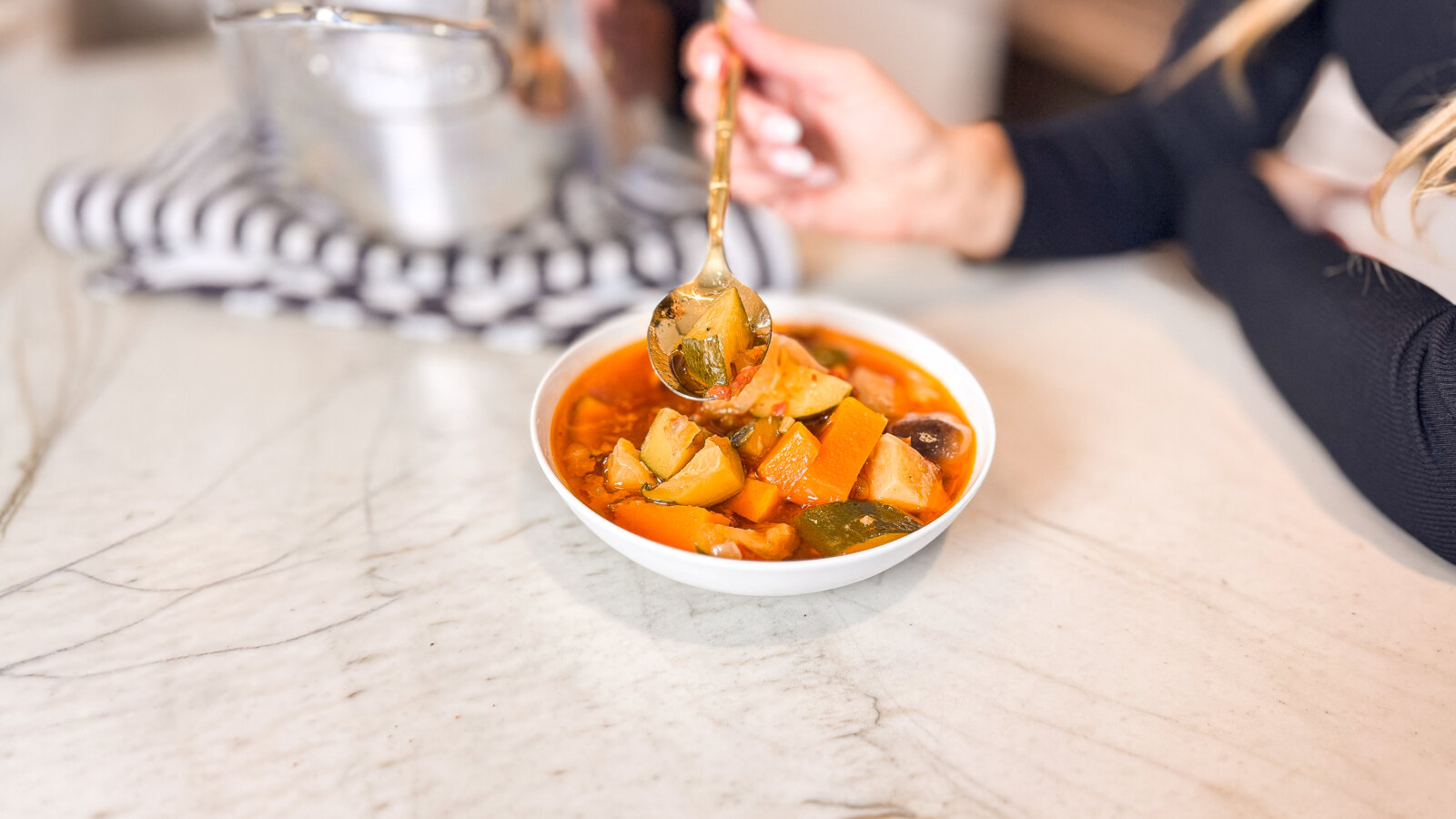 A bowl of hearty beef and vegetable soup with chunks of pumpkin, zucchini, mushrooms, and root vegetables, held by a gold spoon over a marble countertop.