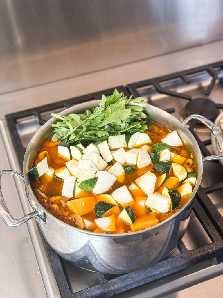 A large stainless steel pot of beef and vegetable soup simmering on the stove, filled with zucchini, turnip, pumpkin, and topped with fresh herbs before stirring.