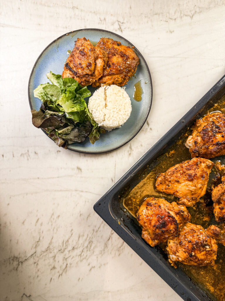 Overhead view of a plate with spicy baked chicken, white rice, and salad greens beside a tray of freshly cooked chicken pieces in sauce.